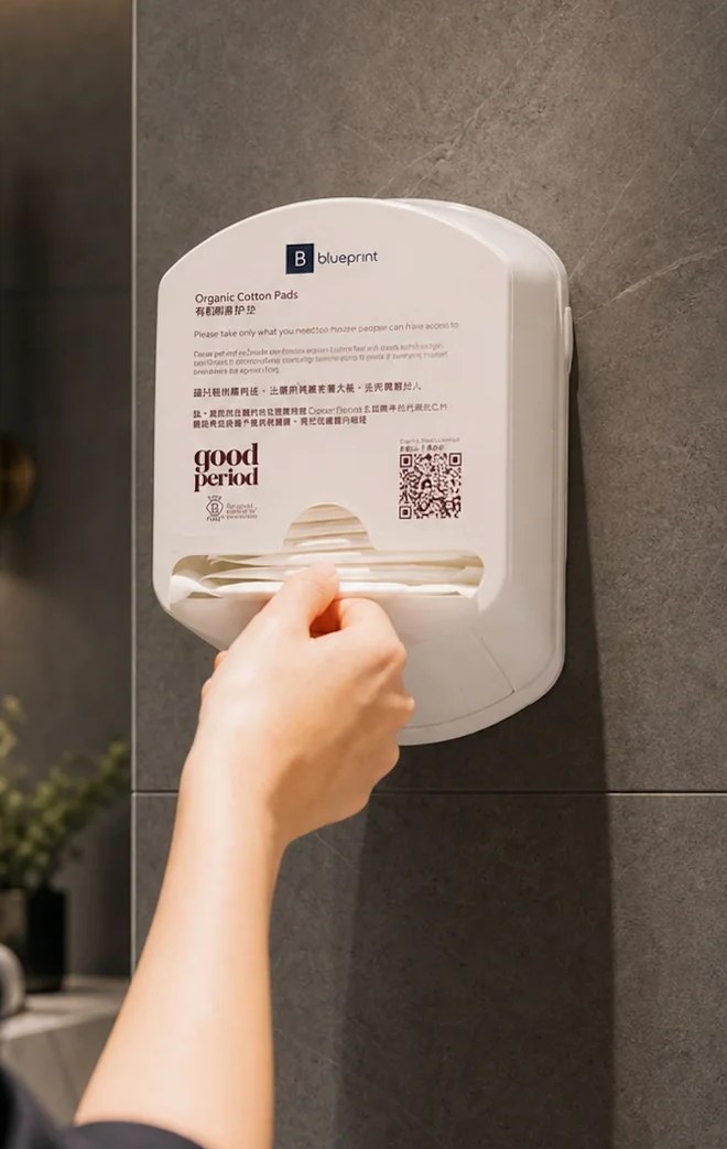 A GoodPeriod wall dispenser of certified organic cotton pads mounted in a workplace washroom, with a hand taking a pad